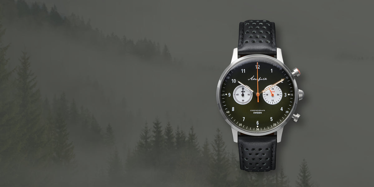 A wristwatch with a black textured strap, silver-toned case, and dark green dial, shown against a misty pine forest background. From Akerfalk watches