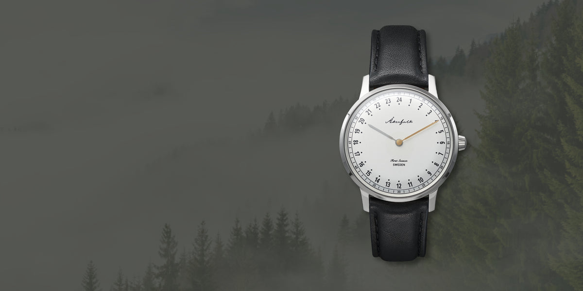 A wristwatch with a black leather strap, silver-toned case, and white dial, shown against a misty forest background. The brand name 'Anchors' is visible on the dial. 24-hour watch from Akerfalk